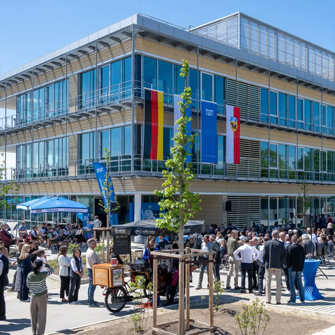 Ein Gebäude mit vielen Fenstern und Glasfassade ist zu sehen. Im Vordergrund stehen viele Menschentrauben vor dem Gebäude, geschart um Stehtische und junge Bäume. An der Fassade hängen die Flaggen Deutschlands, Bayerns, der TUM und der Stadt Freising.