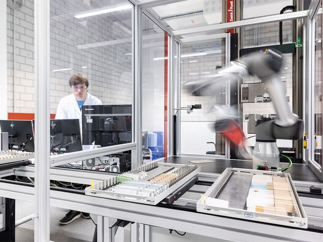 Der Roboterarm befindet sich in einer Einhausung, wo die Probenmagazine über ein Linearschienensystem zugeführt werden. Der Roboterarm befindet sich in einer Einhausung, wo die Probenmagazine über ein Linearschienensystem zugeführt werden.