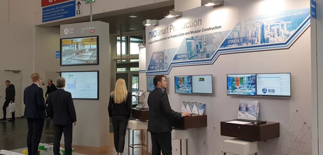 RIB Software präsentiert RIB One Prefab Ein Mann im Anzug steht von den Informations-Monitoren auf dem RIB Software-Stand auf einer Messe