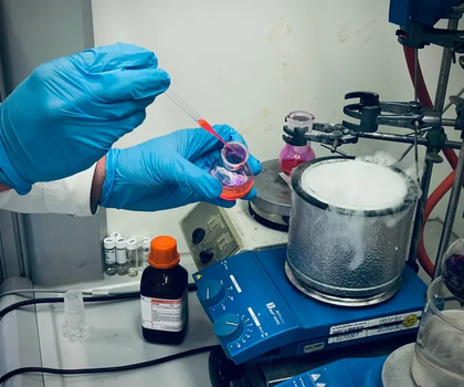 Synthesechemie: Forscher entwickeln neue Alkyl-Swap-Methode für N-Methylamine. Auf dem Bild sind die Hände eines CHemikers mit blauen Handschuhen zu sehen, der mit einer Pipette eine rote Flüssigkeit in ein dampfendes rundes Gefäß auf einer blauen beheizten Rührplatte gibt.