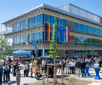 Zentrum für Infektionsprävention: Neuer Forschungsbau an der TUM eröffnet. Das Zentrum für Infektionsprävention (ZIP) steht am Campus Freising-Weihenstephan und ist Teil der TUM School of Life Sciences. Unter Leitung des wissenschaftlichen Direktors Dietmar Zehn wird hier unter anderem zu multiresistenten Keimen geforscht. Ein Gebäude mit vielen Fenstern und Glasfassade ist zu sehen. Im Vordergrund stehen viele Menschentrauben vor dem Gebäude, geschart um Stehtische und junge Bäume. An der Fassade hängen die Flaggen Deutschlands, Bayerns, der TUM und der Stadt Freising.
