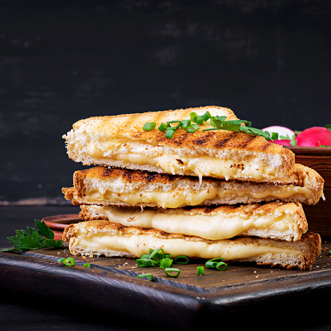 Geröstetes Toas mit geschmolzenem Käse auf einem Holztisch vor schwarzem Hintergrund. Hinten rechts eine Schale mit Käutern und Radieschen.