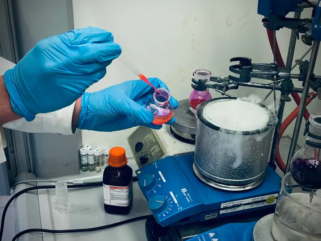 Chemikerinnen und Chemiker der Universität Wien entwickeln eine Methode zur gezielten Modifikation von N-Methylaminen mit Potenzial für die Wirkstoffforschung. Synthesechemie: Forscher entwickeln neue Alkyl-Swap-Methode für N-Methylamine. Auf dem Bild sind die Hände eines CHemikers mit blauen Handschuhen zu sehen, der mit einer Pipette eine rote Flüssigkeit in ein dampfendes rundes Gefäß auf einer blauen beheizten Rührplatte gibt.