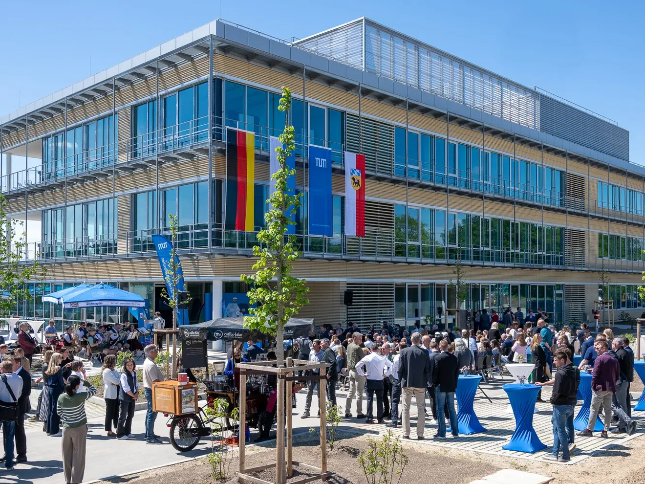 Ein Gebäude mit vielen Fenstern und Glasfassade ist zu sehen. Im Vordergrund stehen viele Menschentrauben vor dem Gebäude, geschart um Stehtische und junge Bäume. An der Fassade hängen die Flaggen Deutschlands, Bayerns, der TUM und der Stadt Freising.