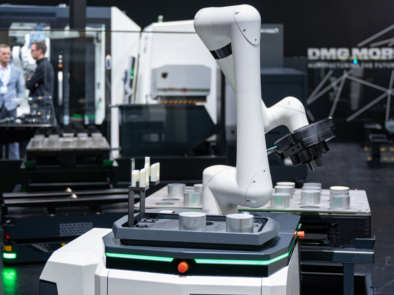 Ein kollaborativer Industrieroboter auf mobiler Plattform bewegt Werkstücke in einer Produktionsumgebung; im Hintergrund sind CNC-Maschinen und zwei Personen zu sehen.