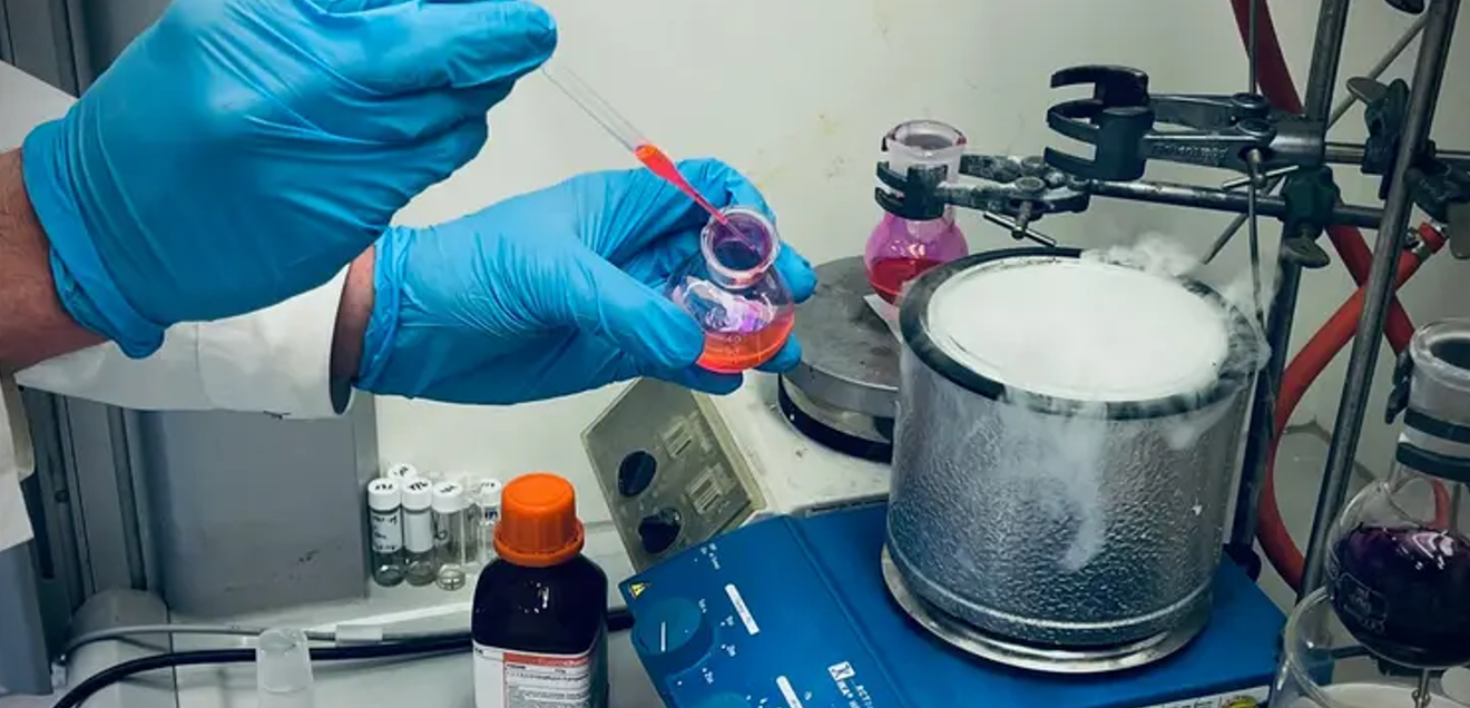 Chemikerinnen und Chemiker der Universität Wien entwickeln eine Methode zur gezielten Modifikation von N-Methylaminen mit Potenzial für die Wirkstoffforschung. Synthesechemie: Forscher entwickeln neue Alkyl-Swap-Methode für N-Methylamine. Auf dem Bild sind die Hände eines CHemikers mit blauen Handschuhen zu sehen, der mit einer Pipette eine rote Flüssigkeit in ein dampfendes rundes Gefäß auf einer blauen beheizten Rührplatte gibt.