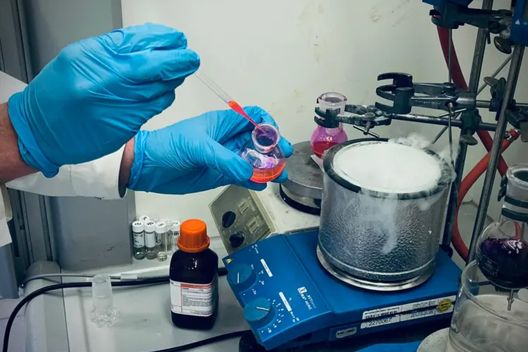 Chemikerinnen und Chemiker der Universität Wien entwickeln eine Methode zur gezielten Modifikation von N-Methylaminen mit Potenzial für die Wirkstoffforschung. Synthesechemie: Forscher entwickeln neue Alkyl-Swap-Methode für N-Methylamine. Auf dem Bild sind die Hände eines CHemikers mit blauen Handschuhen zu sehen, der mit einer Pipette eine rote Flüssigkeit in ein dampfendes rundes Gefäß auf einer blauen beheizten Rührplatte gibt.