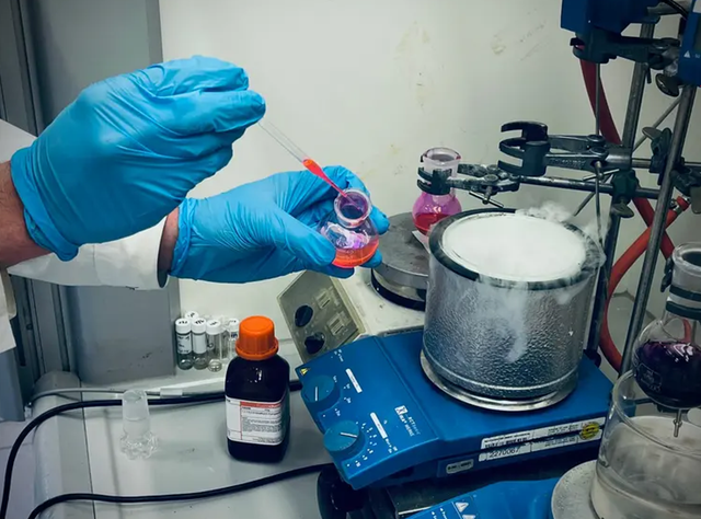 Chemikerinnen und Chemiker der Universität Wien entwickeln eine Methode zur gezielten Modifikation von N-Methylaminen mit Potenzial für die Wirkstoffforschung. Synthesechemie: Forscher entwickeln neue Alkyl-Swap-Methode für N-Methylamine. Auf dem Bild sind die Hände eines CHemikers mit blauen Handschuhen zu sehen, der mit einer Pipette eine rote Flüssigkeit in ein dampfendes rundes Gefäß auf einer blauen beheizten Rührplatte gibt.