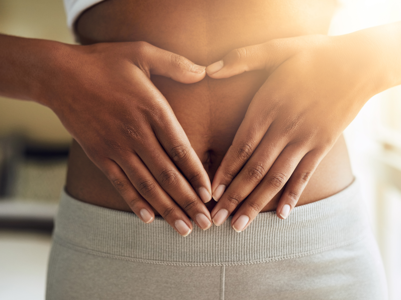 Darmmikrobiom: Studie zeigt Einfluss aufgenommener Mikroorganismen aus der Nahrung Eine Frau hat die Hände zum Herz geformt auf höhe des Darms. Im Hintergrund Sonnenstrahlen. Symbolbild für Darmgesundheit durch ein vielfältiges Mikrobiom.