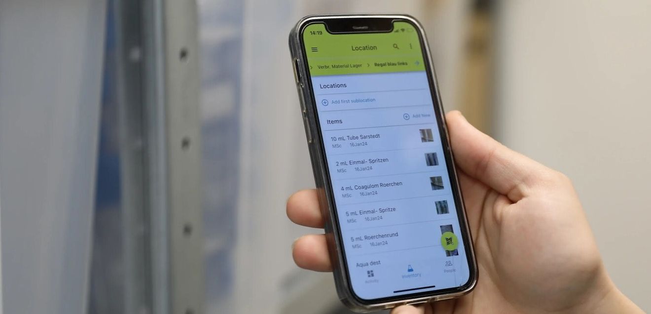Digitalisierung von Laborbeständen und Lagerorten Studierende im Ausbildungslabor hält ein Mobiltelefon in der Hand, auf dem Lagerbestände der Labor-Materialien und Bestände der Chemikalien im Labor angezeigt werden.