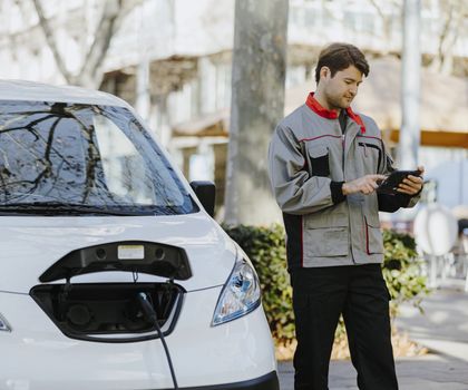 Webfleet ist neuer Netzwerkpartner von AllianZ-SmartUrbanLogistics Ein Mann steht vor einem kleinem, weißen E-Auto, das gerade geladen wird. Dabei prüft er Daten auf einem kleinen Tablet.