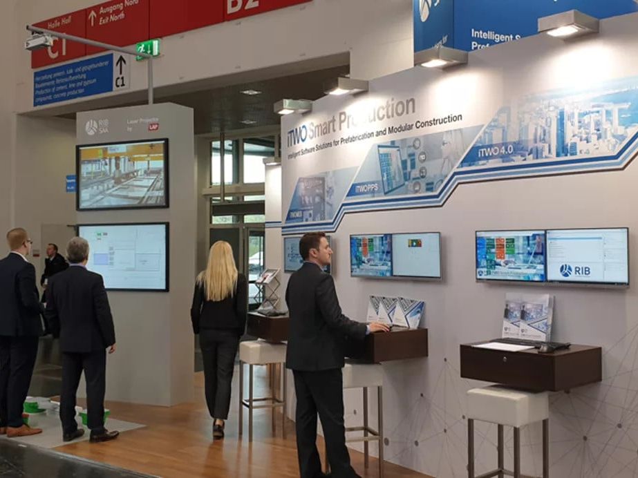 Ein Mann im Anzug steht von den Informations-Monitoren auf dem RIB Software-Stand auf einer Messe