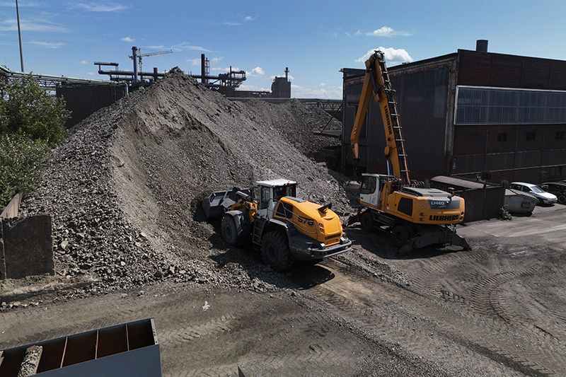Bei der Firma Heis sind mehrere Erdbewegungsmaschinen von Liebherr im Einsatz. Ein Liebherr Radlader nimmt mit seiner Schaufel Material auf, daneben steht ein Liebherr Raupenbagger