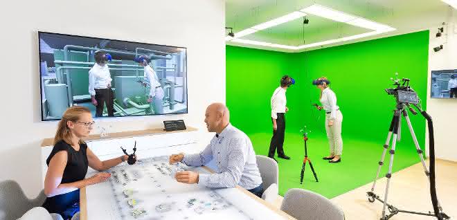 Virtuelle Realität im „Autodesk VR Lab“ München: Die Personen in der Greenbox „begehen“ das BIM-Modell mittels VR-Brille und begutachten so u. a. Wege und Flächen. Gleichzeitig werden die beiden von außen beraten und geleitet. So können Fachplaner, Nutzer und Sachverständige (hier für Laborabnahme) das Gebäude in der Planung bewerten und optimieren, wodurch sich kostenintensive Änderungen in späteren Phasen vermeiden lassen. Virtuelle Realität im „Autodesk VR Lab“ München: 2 Personen stehend mit VR-Brillen, auf einem Bildschirm an der Wand ein virtuelles Bild, und 2 Personen sitzen am Schreibtisch.
