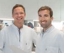 Krebs-Immuntherapien, die auf sogenannte Checkpoint-Inhibitoren setzen, schlagen deutlich besser an, wenn in den Tumorzellen das Protein RIG-I aktiv ist. Das konnten PD Dr. Hendrik Poeck (links) und PD Dr. Simon Heidegger (r.) in einer aktuellen Studie zeigen – ein möglicher Ansatz für zukünftige Therapien. PD Dr. Hendrik Poeck und PD Dr. Simon Heidegger stehen in einem Labor, beide in weißen Kitteln.