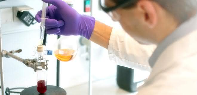 Forschende der TU Graz und Uni Wien stellen eine neue Methode zur Herstellung lipidierter Proteine vor, die einfach durchzuführen und flexibel anwendbar ist. Im Labor: Mann im Kittel gibt mit Hilfe einer Spritze eine farblose Flüssigkeit in einen Glaskolben, der eine rote Flüssigkeit enthält.