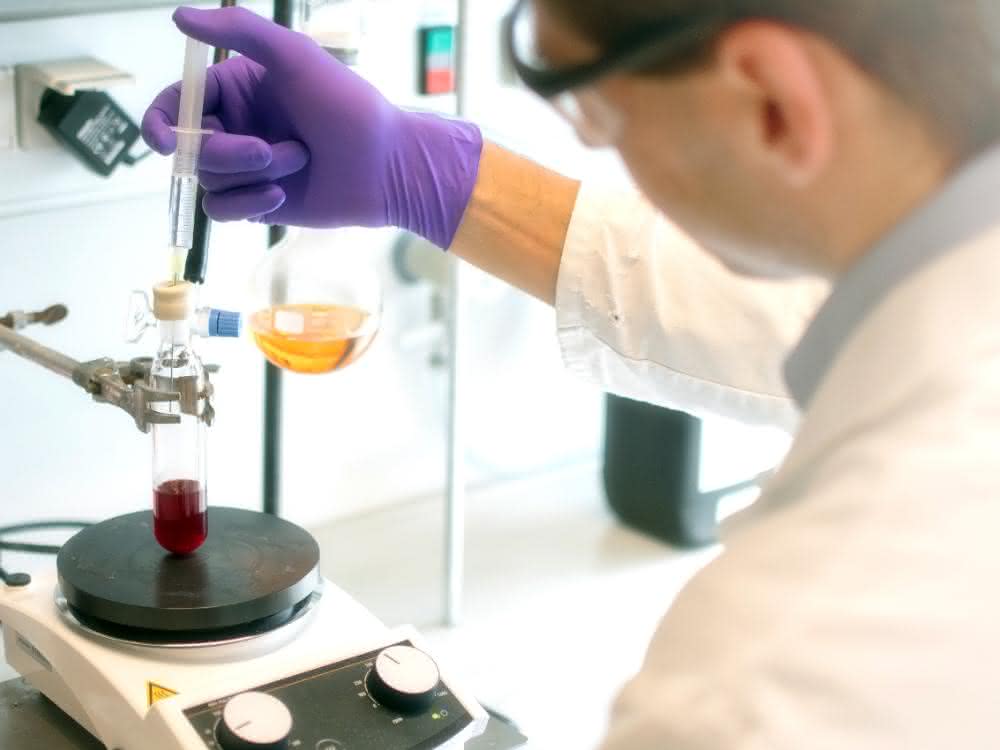Forschende der TU Graz und Uni Wien stellen eine neue Methode zur Herstellung lipidierter Proteine vor, die einfach durchzuführen und flexibel anwendbar ist. Im Labor: Mann im Kittel gibt mit Hilfe einer Spritze eine farblose Flüssigkeit in einen Glaskolben, der eine rote Flüssigkeit enthält.
