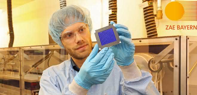 Dr. Andreas Distler (ZAE Bayern) mit dem organischen Rekord-Solarmodul in der Solarfabrik der Zukunft neben der Pilotlinie für Dünnschicht-Photovoltaik. Ein Mann, bekleidet mit Kittel, Haarnetz und Handschuhen hält das Photovolatikmodul (kleine Platte) in die Höhe und betrachtet sie.
