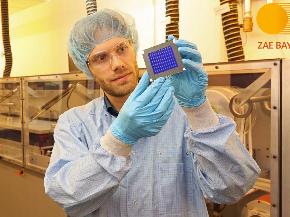 Dr. Andreas Distler (ZAE Bayern) mit dem organischen Rekord-Solarmodul in der Solarfabrik der Zukunft neben der Pilotlinie für Dünnschicht-Photovoltaik. Ein Mann, bekleidet mit Kittel, Haarnetz und Handschuhen hält das Photovolatikmodul (kleine Platte) in die Höhe und betrachtet sie.