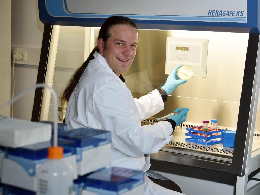 Prof. Kogler (im Laborkittel und mit Handschuhen) vor einer Workstation, in der er eine Petrischale hochhält.