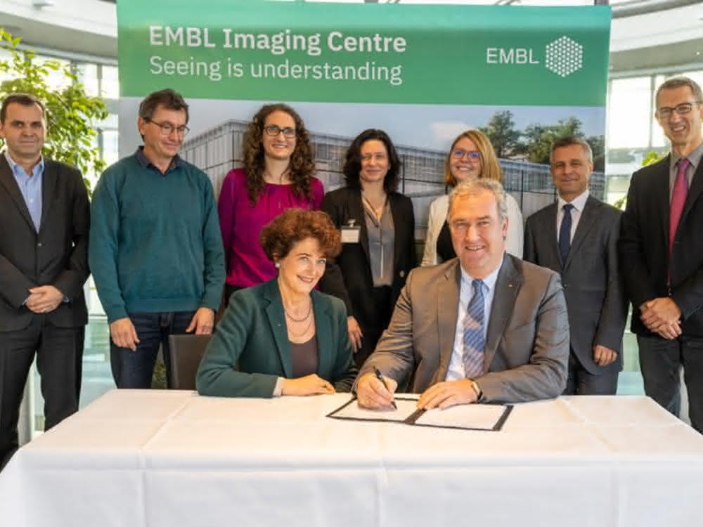 Gruppenfoto von Vertretern von Leica Microsystems und vom EMBL; im Vordergrund am Tisch sitzend Edith Hard und Markus Lusser bei der Unterzeichnung der Vereinbarung