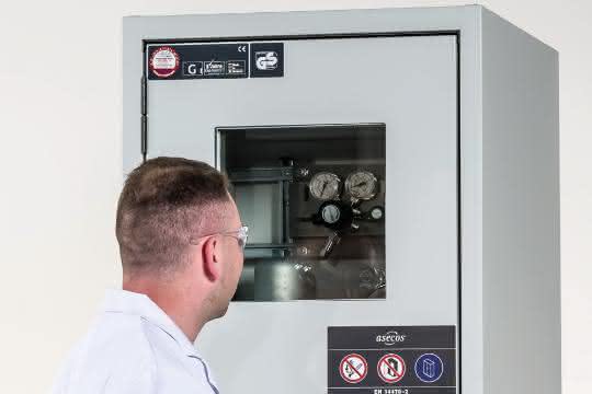 Mann im Kittel blickt durch das Türfenster des Druckgasflaschenschranks